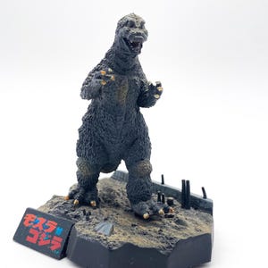 Peut inclure: Figurine de Godzilla dans une pose menaçante, sur un socle de diorama détaillé. La figurine est principalement gris foncé avec des accents jaunes sur ses griffes et ses genoux. La base présente une surface texturée et une étiquette de texte japonaise.