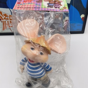 Może przedstawiać: Lalka Soft Topo Gigio w przezroczystym plastikowym opakowaniu. Lalka ma duże różowe uszy, słomkowy kapelusz i niebiesko-białą koszulę w paski. Opakowanie zawiera japoński tekst i ilustracje postaci.