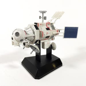 Op de afbeelding: Gedetailleerd model van een ruimteschip, voornamelijk wit met rode accenten, gemonteerd op een zwarte basis. Het heeft zonnepanelen, antennes en een bolvormig voorste gedeelte. Het model staat tegen een witte achtergrond.