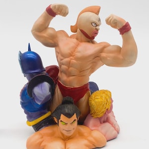 May include: A detailed figurine featuring several muscular figures in various poses. The central figure flexes its biceps, wearing red wristbands and briefs. Other figures include a sumo wrestler and a character in a blue helmet.
