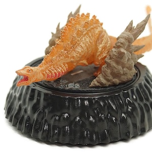 May include: A detailed figurine of a dinosaur-like creature in shades of orange and brown, with a red mouth, atop a black, textured base. The creature has a spiky back and tail, and is posed as if in motion.