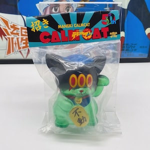 May include: A Maneki Calmcat figurine in a clear plastic bag. The cat is green with a black head and ears, and has red eyes and a gold coin. The packaging has Japanese text and the words "CALMCAT".