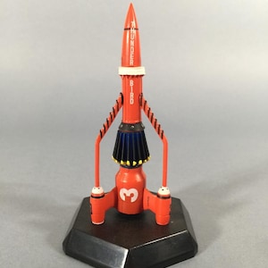 Puede incluir: Un modelo de cohete rojo con el texto "Thunderbird Bird". El cohete tiene una banda blanca, detalles azules y amarillos y el número "3". Está montado sobre una base hexagonal negra, un juguete clásico.
