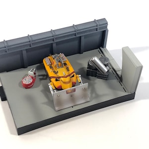Pode incluir: Diorama em miniatura com um veículo amarelo e laranja com uma lâmina prateada, uma estrutura vermelha e branca e um cilindro prateado. A cena é montada numa plataforma cinzenta com paredes cinzentas.