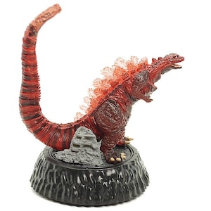 May include: A red and black Godzilla figurine with a long, curved tail and spiky dorsal fins. The figurine is mounted on a black, textured base with a small rock formation. The overall design is detailed and dynamic.