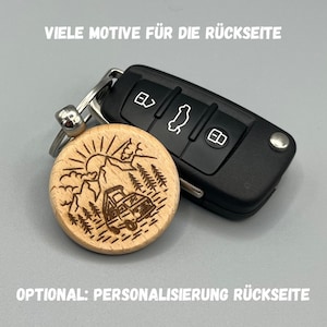 Könnte beinhalten: Ein Holzschlüsselanhänger mit Berg- und Wohnwagendesign, befestigt an einem Autoschlüssel. Der runde Schlüsselanhänger hat eine natürliche Holzoberfläche. Der Autoschlüssel ist schwarz mit weißen Symbolen. Text auf dem Bild lautet: "VIELE MOTIVE FÜR DIE RÜCKSEITE" und "OPTIONAL: PERSONALISIERUNG RÜCKSEITE."