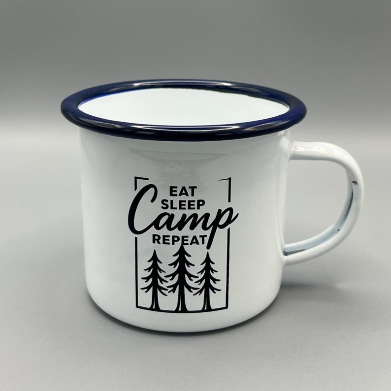 May include: White enamel camping mug with a dark blue rim and handle. The mug features the words "Eat Sleep Camp Repeat" in black, along with a graphic of three pine trees. A classic camping accessory.