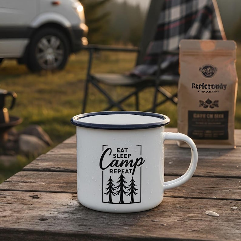 May include: White enamel camping mug with a black rim and handle. The mug features the text "EAT SLEEP CAMP REPEAT" with a graphic of three trees. A bag of coffee sits behind the mug on a wooden table.