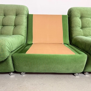 May include: A modular green corduroy sofa with a central tan seat and back. The sofa is composed of three sections, with the outer two featuring tufted cushions and the center section having a flat seat and back. The sofa sits on small, silver-colored feet.