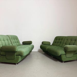 May include: Two green vintage sofas with rounded arms and tufted seat cushions. The sofas are placed side-by-side against a white wall. The sofas have a low profile and are on small, silver casters.