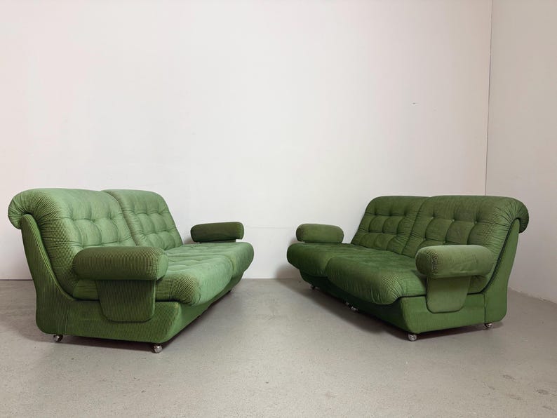 May include: Two green vintage sofas with rounded arms and tufted seat cushions. The sofas are placed side-by-side against a white wall. The sofas have a low profile and are on small, silver casters.