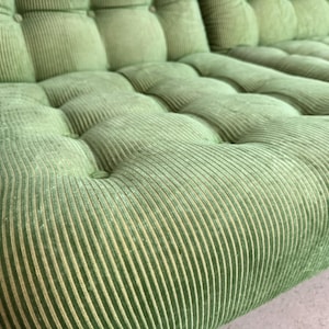 May include: Close-up of a section of a green corduroy sofa. The sofa has a tufted design with button detailing. The fabric has a ribbed texture, and the color is a muted, light green.