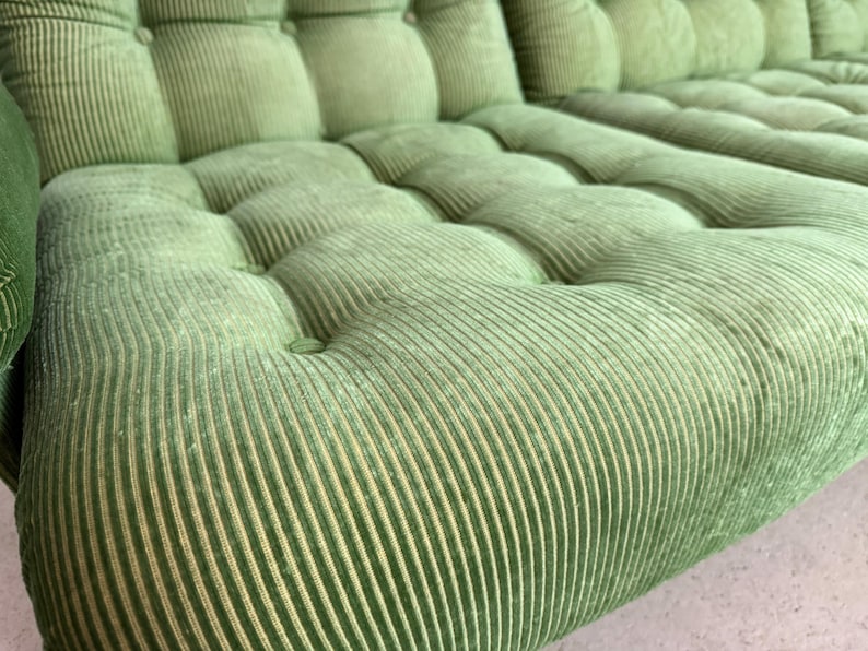 May include: Close-up of a section of a green corduroy sofa. The sofa has a tufted design with button detailing. The fabric has a ribbed texture, and the color is a muted, light green.