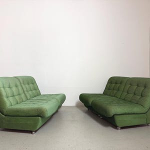 May include: Two green, mid-century modern sofas. Each sofa has a rounded back and armrests, with a tufted seat and back. The sofas are upholstered in a textured green fabric and sit on small, silver casters.