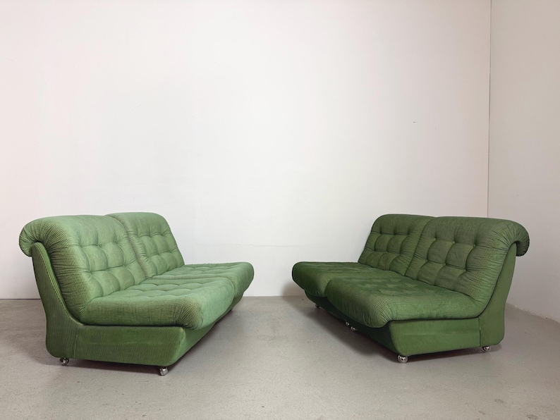 May include: Two green, mid-century modern sofas. Each sofa has a rounded back and armrests, with a tufted seat and back. The sofas are upholstered in a textured green fabric and sit on small, silver casters.