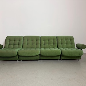 May include: A four-seat, green, tufted sofa with rounded armrests. The sofa has a low profile and sits on small, silver casters. The cushions are square-stitched, and the fabric has a soft, textured appearance. The sofa is against a white wall.