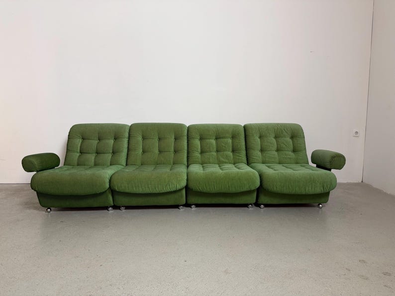 May include: A four-seat, green, tufted sofa with rounded armrests. The sofa has a low profile and sits on small, silver casters. The cushions are square-stitched, and the fabric has a soft, textured appearance. The sofa is against a white wall.