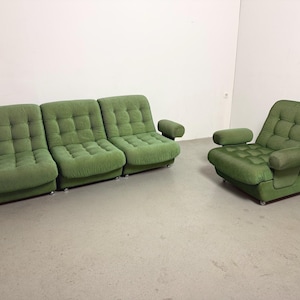 May include: A green upholstered sofa with three sections and a matching armchair. Both pieces have a tufted design and rounded armrests. The furniture is set against a white wall and a light gray floor.