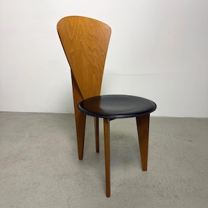 Peut inclure: Une chaise en bois dotée d'un dossier haut et incurvé au grain de bois brun clair, qui s'effile vers le bas en un support central unique. La chaise possède une assise circulaire noire lisse et repose sur trois pieds en bois droit.