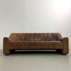 May include: A brown leather sofa with a low profile and a rectangular shape. The sofa has a series of vertical, padded sections across the back and a solid base. The arms are wide and square.