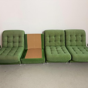 May include: A modular green sofa with a tufted design. The sofa is composed of several sections, including armrests and individual seating units. The fabric has a textured appearance, and the sofa sits on low, silver-colored feet.