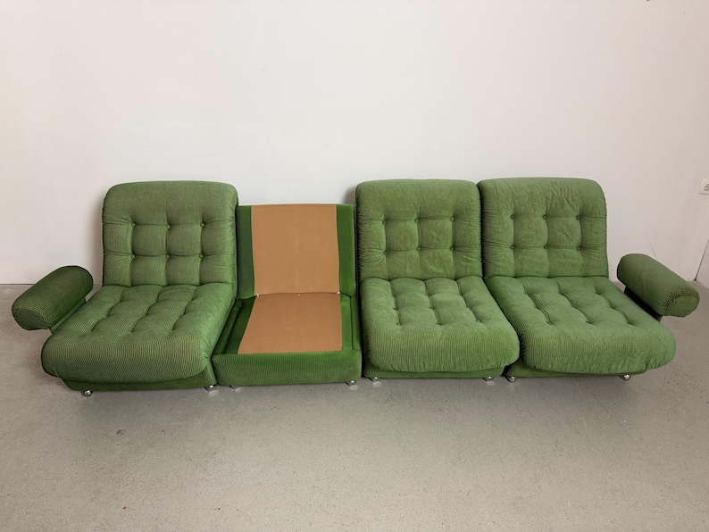 May include: A modular green sofa with a tufted design. The sofa is composed of several sections, including armrests and individual seating units. The fabric has a textured appearance, and the sofa sits on low, silver-colored feet.
