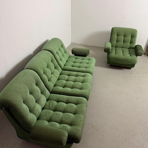 May include: A green corduroy sectional sofa with a matching armchair. The sofa has a rounded corner and a separate headrest. The armchair has armrests. Both pieces have a button-tufted design. The furniture is in a room with a white wall and a concrete floor.