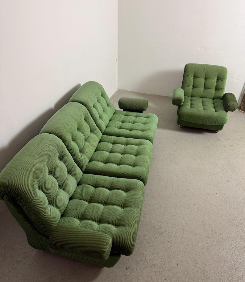 May include: A green corduroy sectional sofa with a matching armchair. The sofa has a rounded corner and a separate headrest. The armchair has armrests. Both pieces have a button-tufted design. The furniture is in a room with a white wall and a concrete floor.