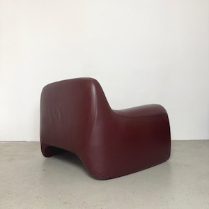 K&ouml;nnte beinhalten: Ein niedriger Sessel, bezogen mit tiefburgunderfarbenem Leder. Der Stuhl zeichnet sich durch eine durchgehende, abgerundete Silhouette mit einer geschwungenen R&uuml;ckenlehne aus, die nahtlos in integrierte Armlehnen &uuml;bergeht und eine solide Form bildet.