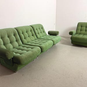 Op de afbeelding: Een groene corduroy bank en fauteuil set. De bank heeft drie secties met gecapitonneerde kussens en afgeronde armleuningen. De fauteuil heeft een vergelijkbaar ontwerp met een vierkante rug en armleuningen. De meubels staan tegen een witte muur.