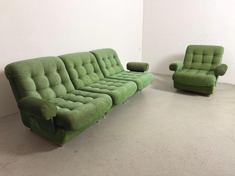 May include: A green corduroy sofa and armchair set. The sofa has three sections with tufted cushions and rounded armrests. The armchair has a similar design with a square back and armrests. The furniture is set against a white wall.