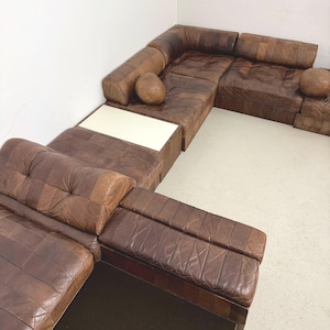 Op de afbeelding: Een modulaire bruin lederen hoekbank met een patchwork ontwerp. De bank heeft een hoekconfiguratie met meerdere secties, waaronder een ingebouwde tafel met een wit oppervlak en twee ronde kussens.