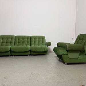 May include: A green, mid-century modern sofa and armchair set. The sofa has three sections with a tufted design, rounded arms, and sits on small silver casters. The matching armchair has a similar design and armrests. The set is in a room with a white wall.