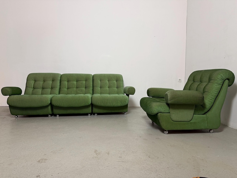 May include: A green, mid-century modern sofa and armchair set. The sofa has three sections with a tufted design, rounded arms, and sits on small silver casters. The matching armchair has a similar design and armrests. The set is in a room with a white wall.