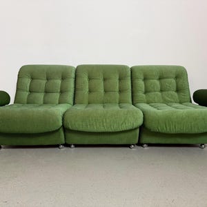 May include: A three-piece green corduroy sofa with rounded armrests. The sofa has a tufted back and seat cushions. The sofa sits on silver-colored, low-profile legs. The overall design is a retro, mid-century modern style.