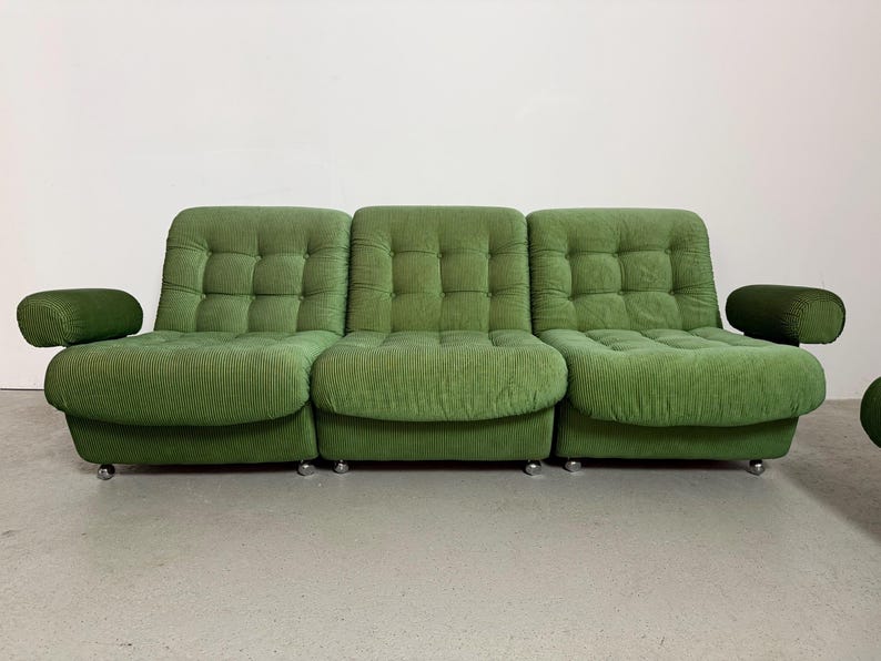 May include: A three-piece green corduroy sofa with rounded armrests. The sofa has a tufted back and seat cushions. The sofa sits on silver-colored, low-profile legs. The overall design is a retro, mid-century modern style.