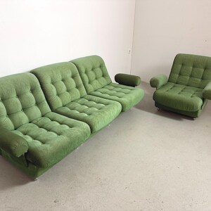 May include: A green, tufted sofa and armchair set. The sofa has three sections with rounded armrests. The armchair has a similar design with armrests. Both pieces are upholstered in a textured fabric and sit on low, silver legs. The set is in a room with white walls and a light-colored floor.