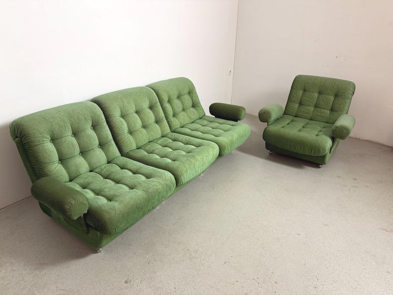 May include: A green, tufted sofa and armchair set. The sofa has three sections with rounded armrests. The armchair has a similar design with armrests. Both pieces are upholstered in a textured fabric and sit on low, silver legs. The set is in a room with white walls and a light-colored floor.