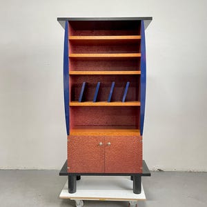 Könnte beinhalten: Ein hoher, farbenfroher Schrank mit schwarzer Ober- und Unterseite. Der Schrank hat einen roten Innenraum mit Holzregalen und blauen Akzenten. Der untere Teil hat zwei Türen mit silbernen Knöpfen. Der Schrank steht auf einer weißen Plattform mit Rädern.