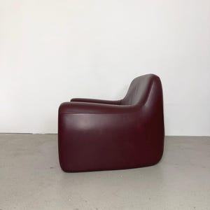 K&ouml;nnte beinhalten: Ein tiefburgunderfarbener Sessel mit einer glatten, durchgehenden Silhouette, abgerundeten Kanten und einem niedrigen Design. Der Sessel ist mit einem einfarbigen Material bezogen und weist einen nahtlosen &Uuml;bergang von der R&uuml;ckenlehne zu den Armlehnen auf.