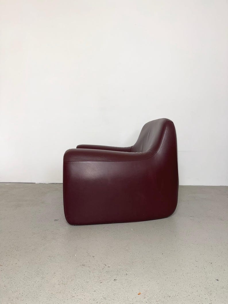 K&ouml;nnte beinhalten: Ein tiefburgunderfarbener Sessel mit einer glatten, durchgehenden Silhouette, abgerundeten Kanten und einem niedrigen Design. Der Sessel ist mit einem einfarbigen Material bezogen und weist einen nahtlosen &Uuml;bergang von der R&uuml;ckenlehne zu den Armlehnen auf.