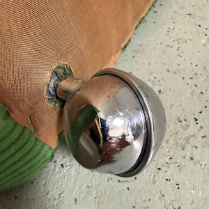 May include: Close-up of a shiny, chrome-colored bell attached to a brown fabric. The bell is round with a visible seam and reflects the surrounding environment. Green fabric is visible on the edge of the brown fabric.