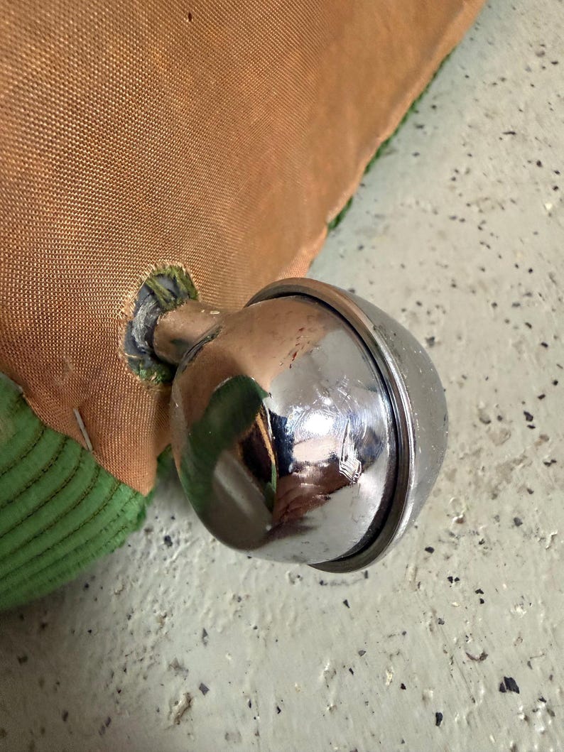 May include: Close-up of a shiny, chrome-colored bell attached to a brown fabric. The bell is round with a visible seam and reflects the surrounding environment. Green fabric is visible on the edge of the brown fabric.