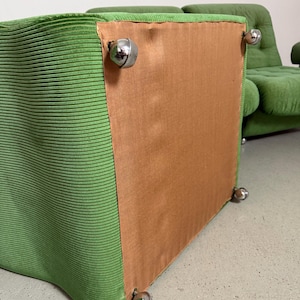 May include: A green corduroy upholstered chair with chrome ball feet. The chair has a boxy shape and a light brown backing. Another chair is visible in the background.