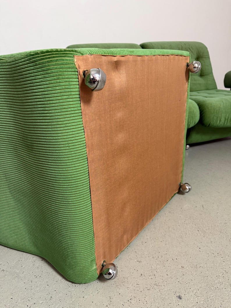 May include: A green corduroy upholstered chair with chrome ball feet. The chair has a boxy shape and a light brown backing. Another chair is visible in the background.