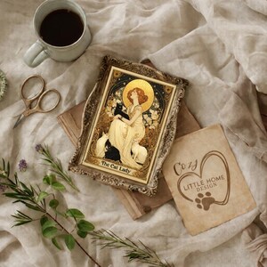 May include: An Art Nouveau style framed print titled "The Cat Lady" featuring a woman with cats, a coffee cup, scissors, and a small dish. The image is set on a beige fabric with sprigs of rosemary and a "Cozy Little Home Design" card.