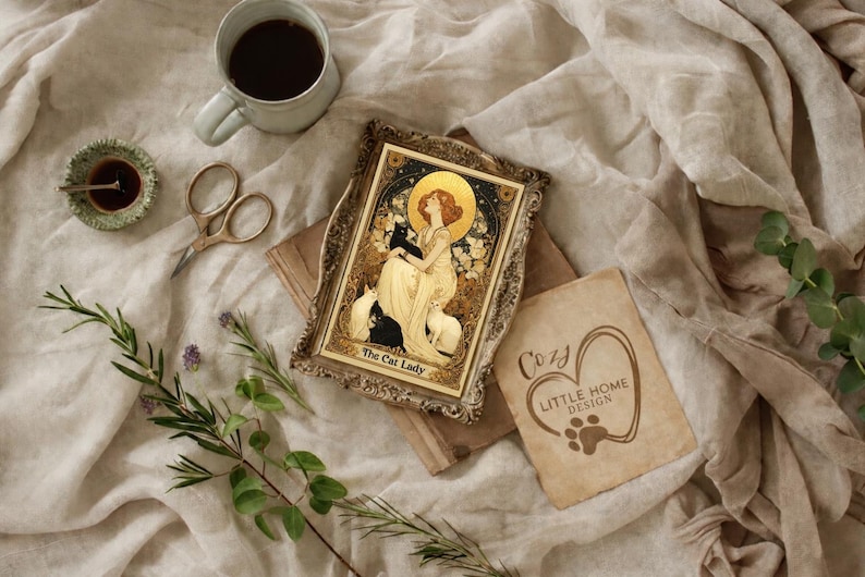May include: An Art Nouveau style framed print titled "The Cat Lady" featuring a woman with cats, a coffee cup, scissors, and a small dish. The image is set on a beige fabric with sprigs of rosemary and a "Cozy Little Home Design" card.