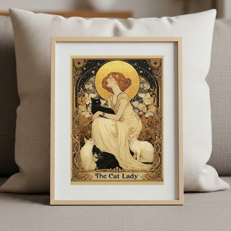May include: A framed art print titled The Cat Lady featuring an illustration of a person in a long cream dress with a golden halo, surrounded by two black cats and two white cats, set within an ornate gold and black decorative border.