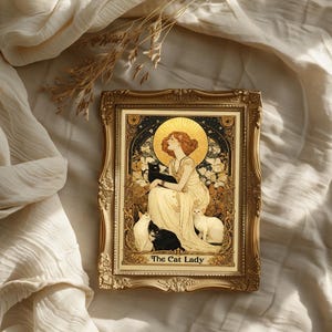 May include: A framed art print featuring a woman with red hair and a golden halo, surrounded by cats. The artwork is in a gold ornate frame, with the text "The Cat Lady" at the bottom. The background is a cream-colored fabric.