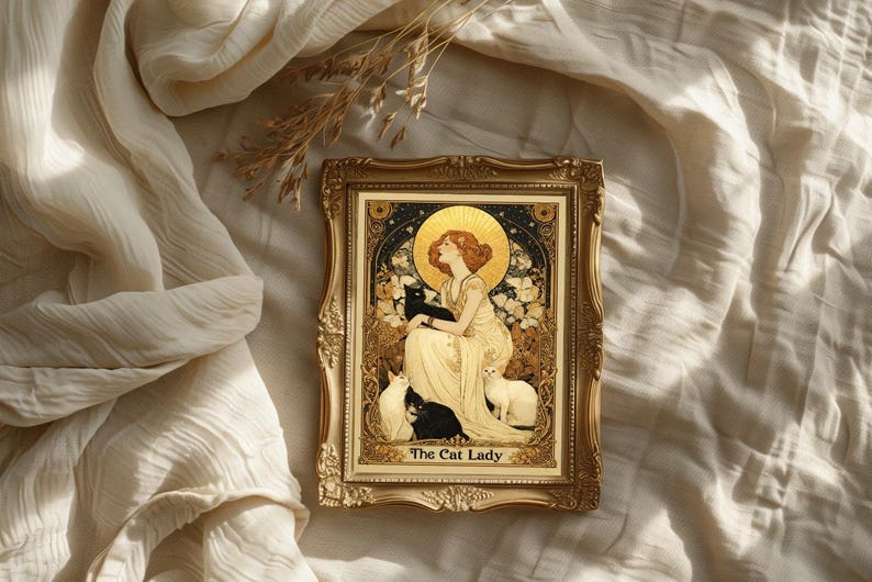 May include: A framed art print featuring a woman with red hair and a golden halo, surrounded by cats. The artwork is in a gold ornate frame, with the text "The Cat Lady" at the bottom. The background is a cream-colored fabric.